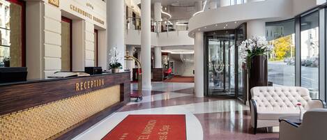 A Grand Hotel lobby with a reception desk, a red carpet with the hotel name, a white sofa, and a large window offering a view of the street.