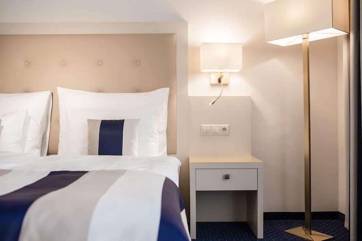A hotel room with a bed, bedside table, lamp, and a wall-mounted light fixture.