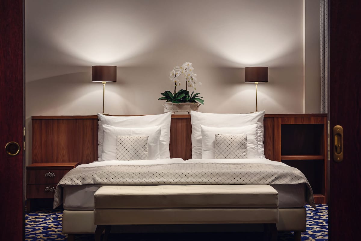 A bedroom with a bed, two bedside lamps, a nightstand, a wooden headboard, a potted plant, and a patterned rug.