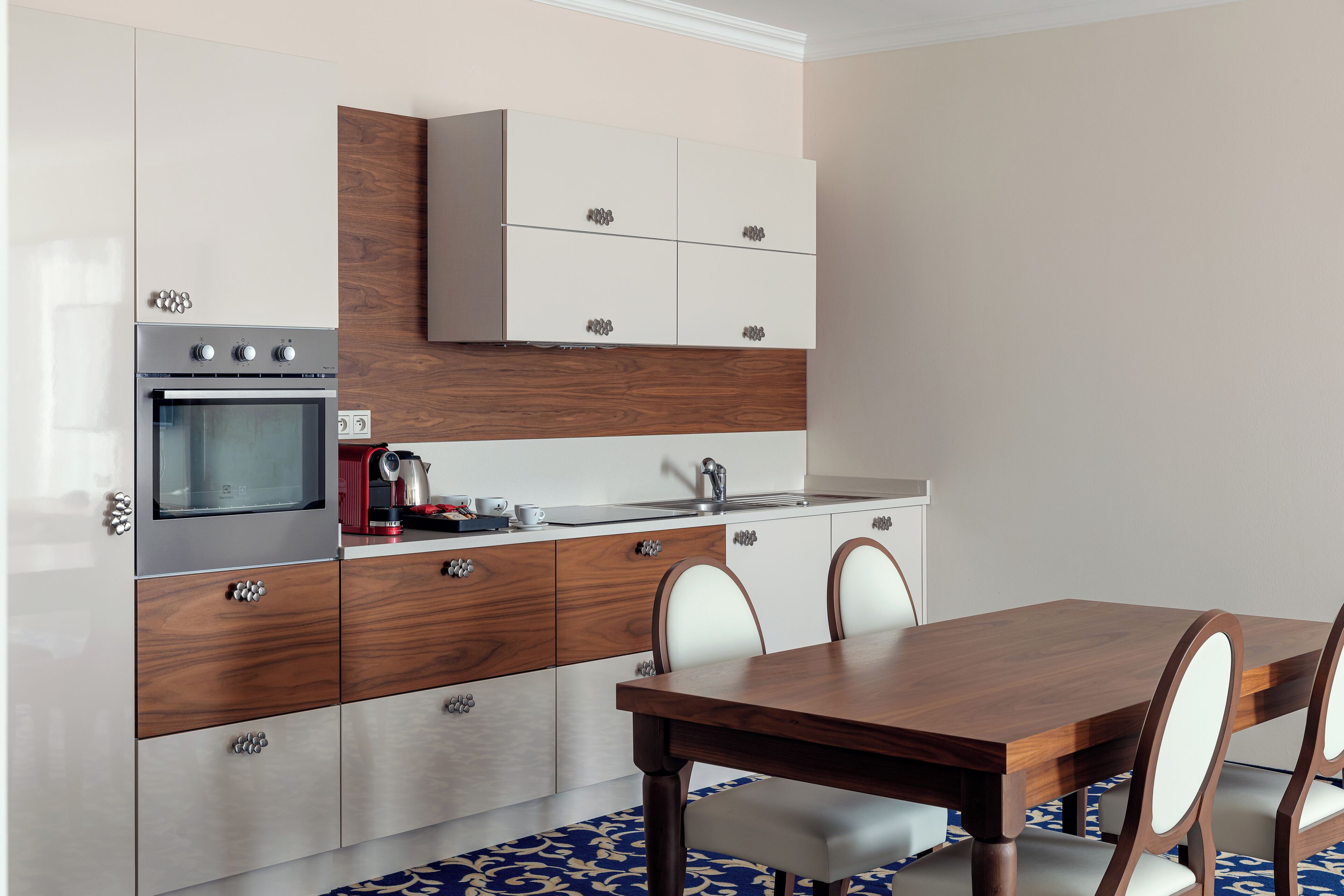 A modern kitchen with a built-in oven, a microwave, and a coffee maker. There is a dining table with chairs and a patterned rug.