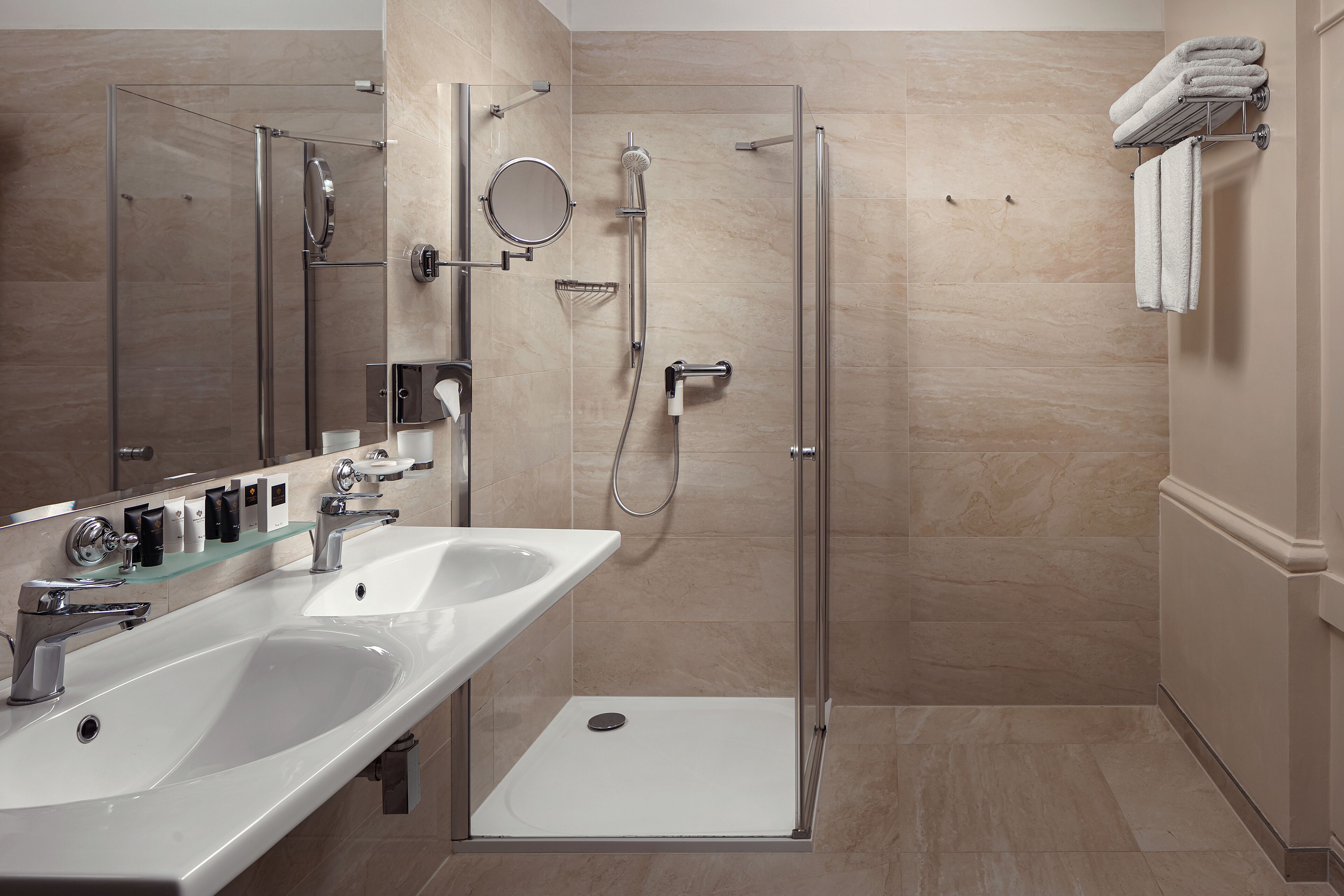 Un baño moderno con ducha de vidrio, dos lavamanos y tocador con artículos de aseo.