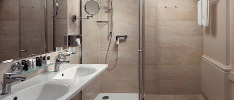 Un baño moderno con ducha de vidrio, dos lavamanos y tocador con artículos de aseo.