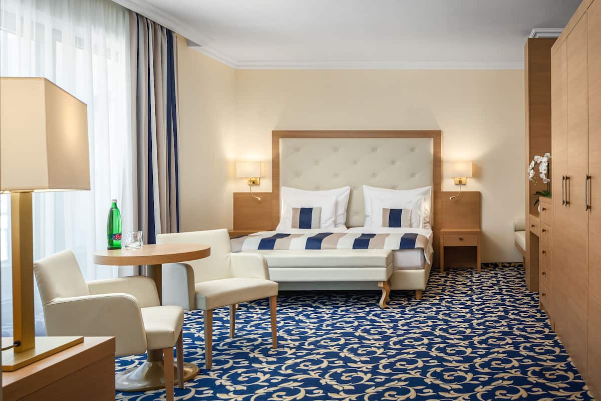 A hotel room with a large bed, two bedside tables, a desk with a chair, a small table, and a patterned carpet.