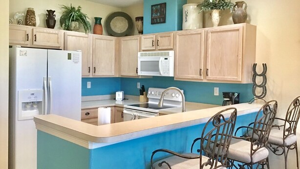 Une cuisine avec un mur d’accent bleu, des armoires en bois, un réfrigérateur blanc, un four à micro-ondes, une cuisinière et un coin bar avec des chaises en métal.