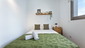 Een slaapkamer met een bed, kussens, een nachtkastje, een wandplank met foto’s en een plant, en een raam.