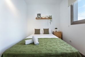 Een slaapkamer met een bed, kussens, een nachtkastje, een wandplank met foto’s en een plant, en een raam.