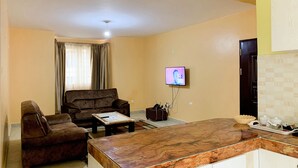 Uma sala de estar com sofá, mesa de centro, TV de tela plana na parede e cozinha com bancada de mármore e armários.