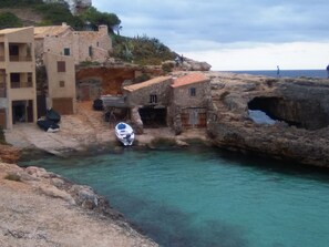 A coastal village with stone buildings, a boat docked near the shore, and clear turquoise water. - Hostal Can Jordi (Santanyi)