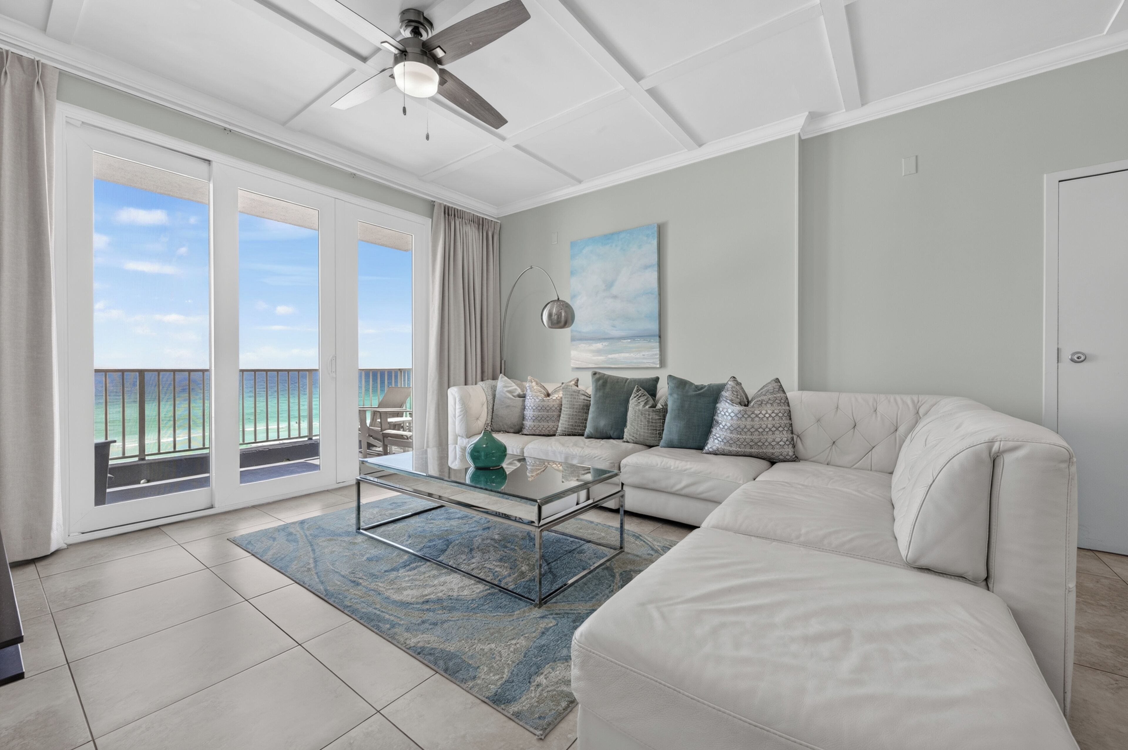 A living room with a white sofa, a glass coffee table, a view of the ocean through large sliding doors, and a ceiling fan.