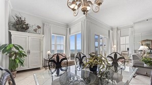 A spacious living room with a glass-top dining table, elegant metal chairs, a chandelier, and large windows offering a view. - Inn at Crystal Beach 210a * Gulf Front with Beach Chair Set. Onsite dining! (Destin)