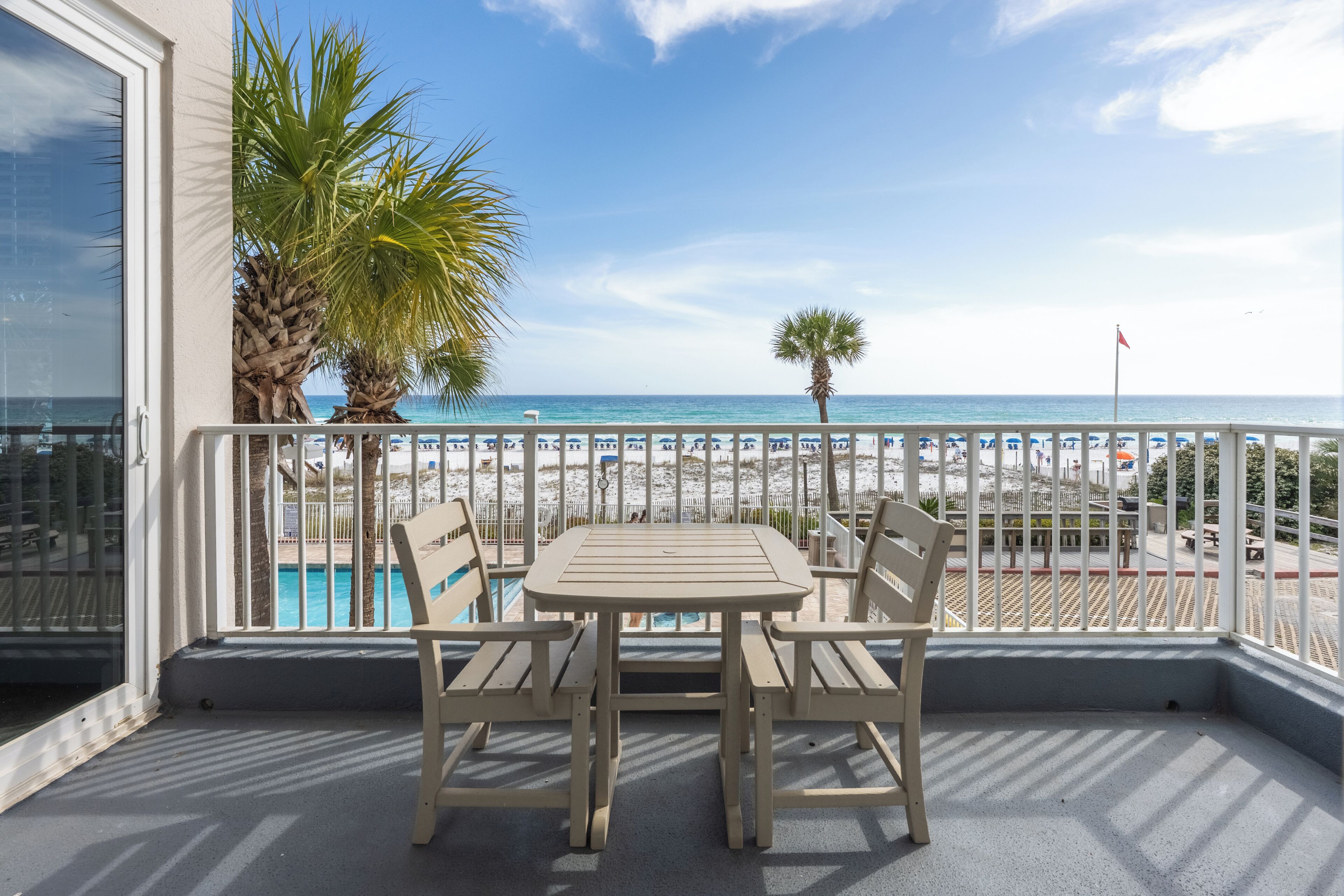 A balcony with a table and chairs, overlooking a beach and pool.