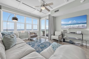 A spacious living room with a large window, a ceiling fan, a flat-screen TV, and a glass coffee table.