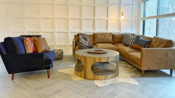 A living room with a dark blue armchair, a light brown sofa, and a round wooden coffee table. - Luxe 10 guest Cabin/Skiing/Pools/Tennis/Gym/Hiking (vernon)