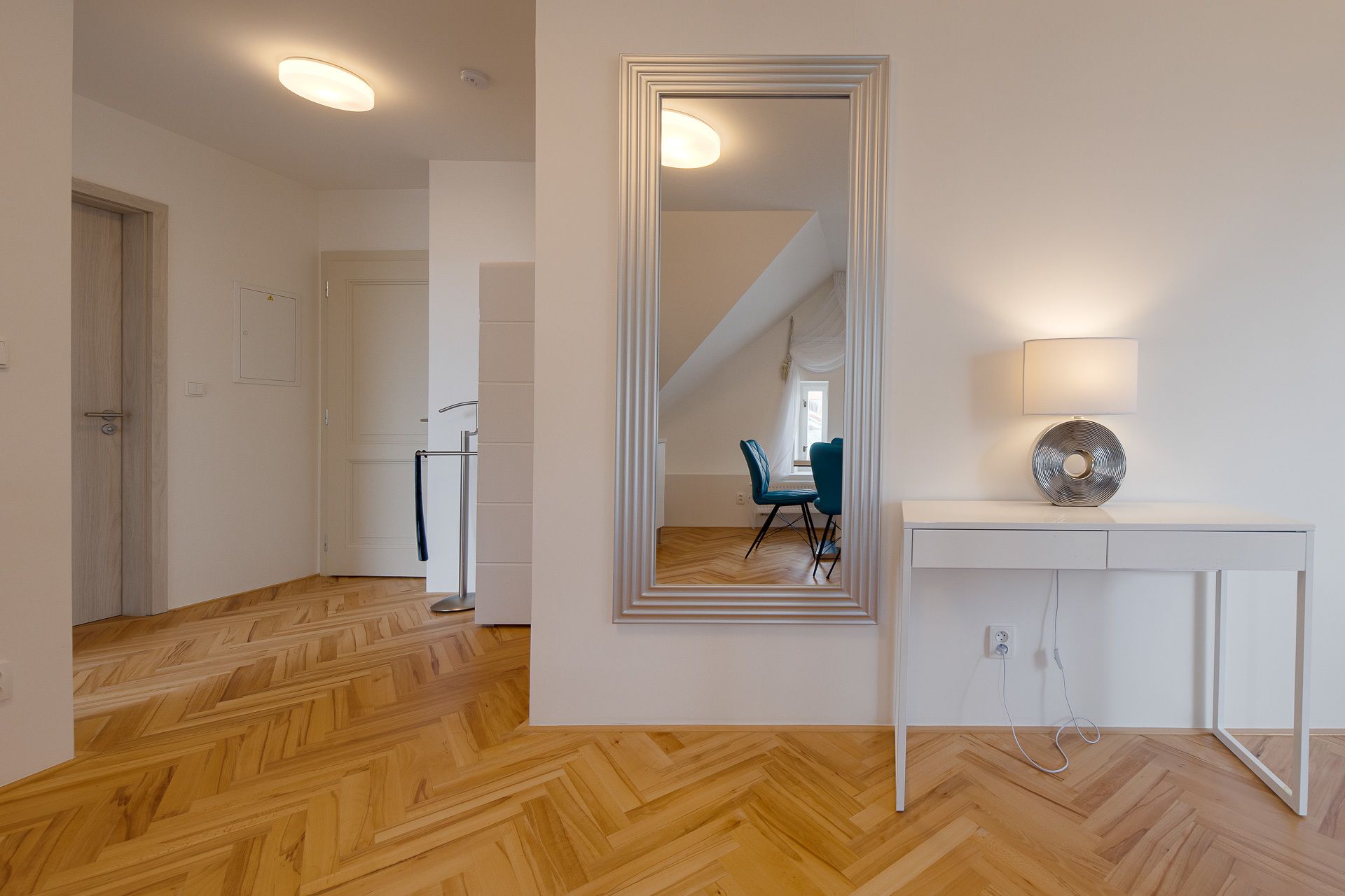 A modern interior with a herringbone wooden floor, a white console table with a lamp, and a large mirror reflecting a room with a blue chair and a window.