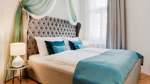 Una camera da letto con una testiera trapuntata, un letto con biancheria bianca e color turchese, un comodino con una lampada e tende trasparenti.