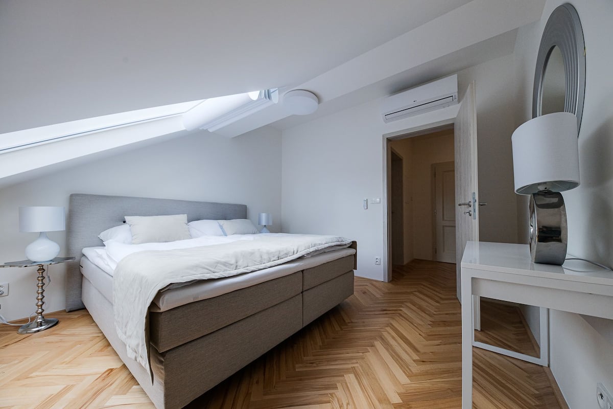 A modern bedroom with a bed, bedside lamps, a ceiling light, and an air conditioning unit.