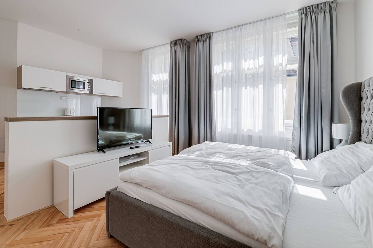 A modern hotel room with a flat-screen TV, a bed with white linens, grey curtains, and a wooden floor.