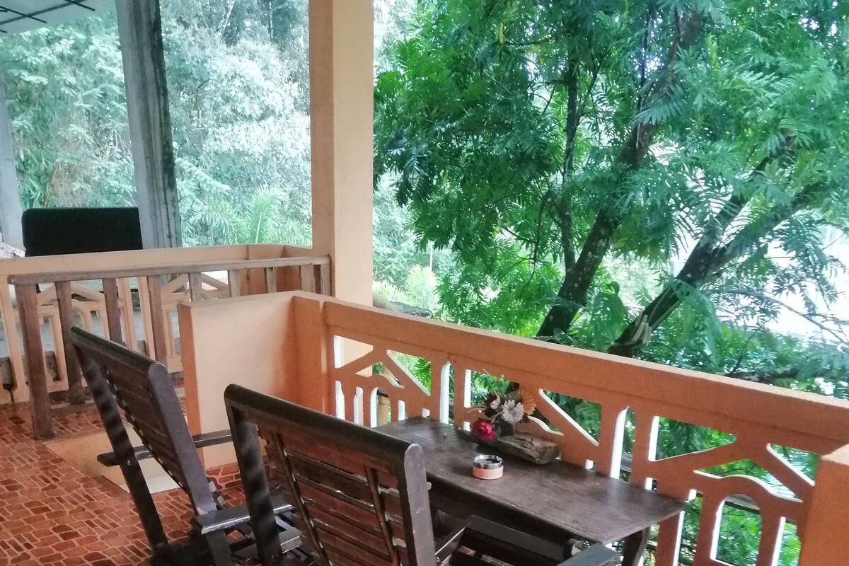 A covered outdoor seating area with a tiled floor, wooden furniture, and a view of lush greenery.