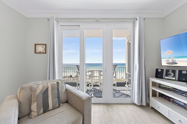 A balcony with a view of the beach, a sofa, a TV, and a bookshelf.