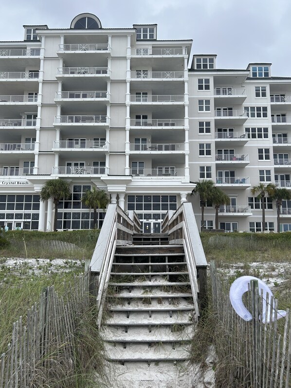 A multi-story hotel with a central staircase leading to the entrance, surrounded by sandy terrain and palm trees.