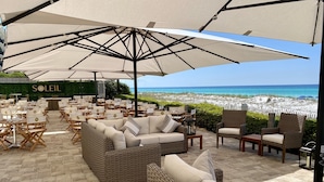 An outdoor seating area with wicker furniture and large umbrellas, overlooking a beach. - Inn at Crystal Beach 103b * Gulf Front with Beach Chair Set. Onsite dining! (Destin)