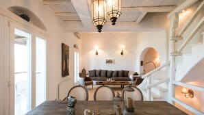A dining area with a wooden table, chairs, and a chandelier. There is a staircase with a railing on the right and a door on the left. - Beautiful Villa at Panormos (Mykonos)