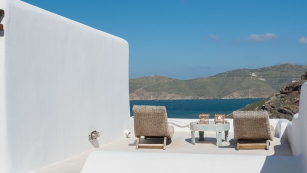 A white-walled terrace with two wicker lounge chairs and a small table, overlooking a coastal landscape with hills and the sea. - Beautiful Villa at Panormos (Mykonos)
