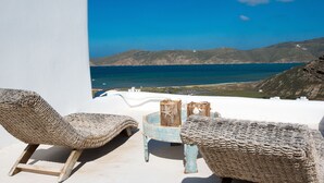A white-walled room with a view of the sea and mountains. There are two woven chairs and a small table with a decorative item. - Beautiful Villa at Panormos (Mykonos)