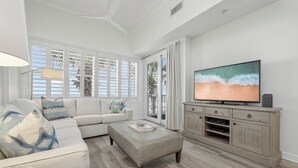 Un salon spacieux avec une grande fenêtre, un ventilateur de plafond, un téléviseur posé sur un meuble en bois et un canapé beige.