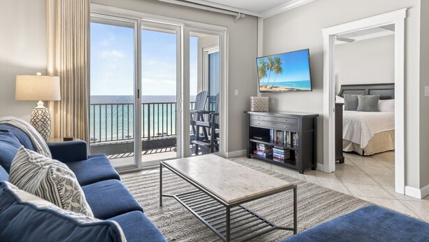 A living room with a sofa, a coffee table, a TV, and a bookshelf. There is a balcony with a view of the ocean. - Inn at Crystal Beach 404 * Gulf Front with Beach Chair Set. Onsite dining! (Destin)