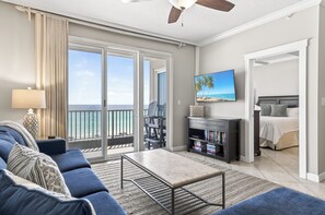 Un salon comprenant un canapé, une table basse, un téléviseur et une bibliothèque. Il y a un balcon avec vue sur l’océan.