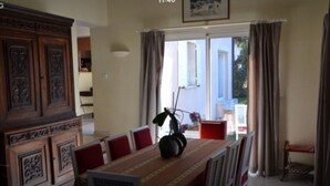 A dining area with a table set for four, a wooden cabinet, and a view of the outside through a window with curtains. - VILLA WITH GARDEN AND CLOSED SWIMMING POOL (Poulx)