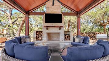 Un patio exterior con chimenea de piedra, asientos con cojines azules y un televisor de pantalla plana sobre el hogar.