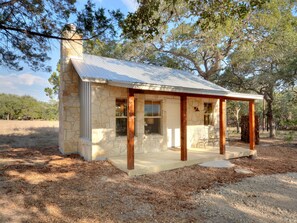 A small stone cabin with a porch, surrounded by trees and a grassy field.