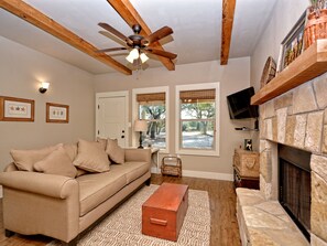 A living room with a beige sofa, a wooden ceiling fan, a stone fireplace, and two windows with blinds.