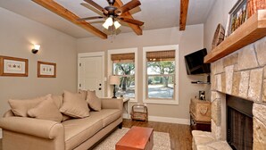 A living room with a beige sofa, a wooden ceiling fan, a stone fireplace, and two windows with blinds. - Texas Sage I Cabin close to the Square! (Wimberley)
