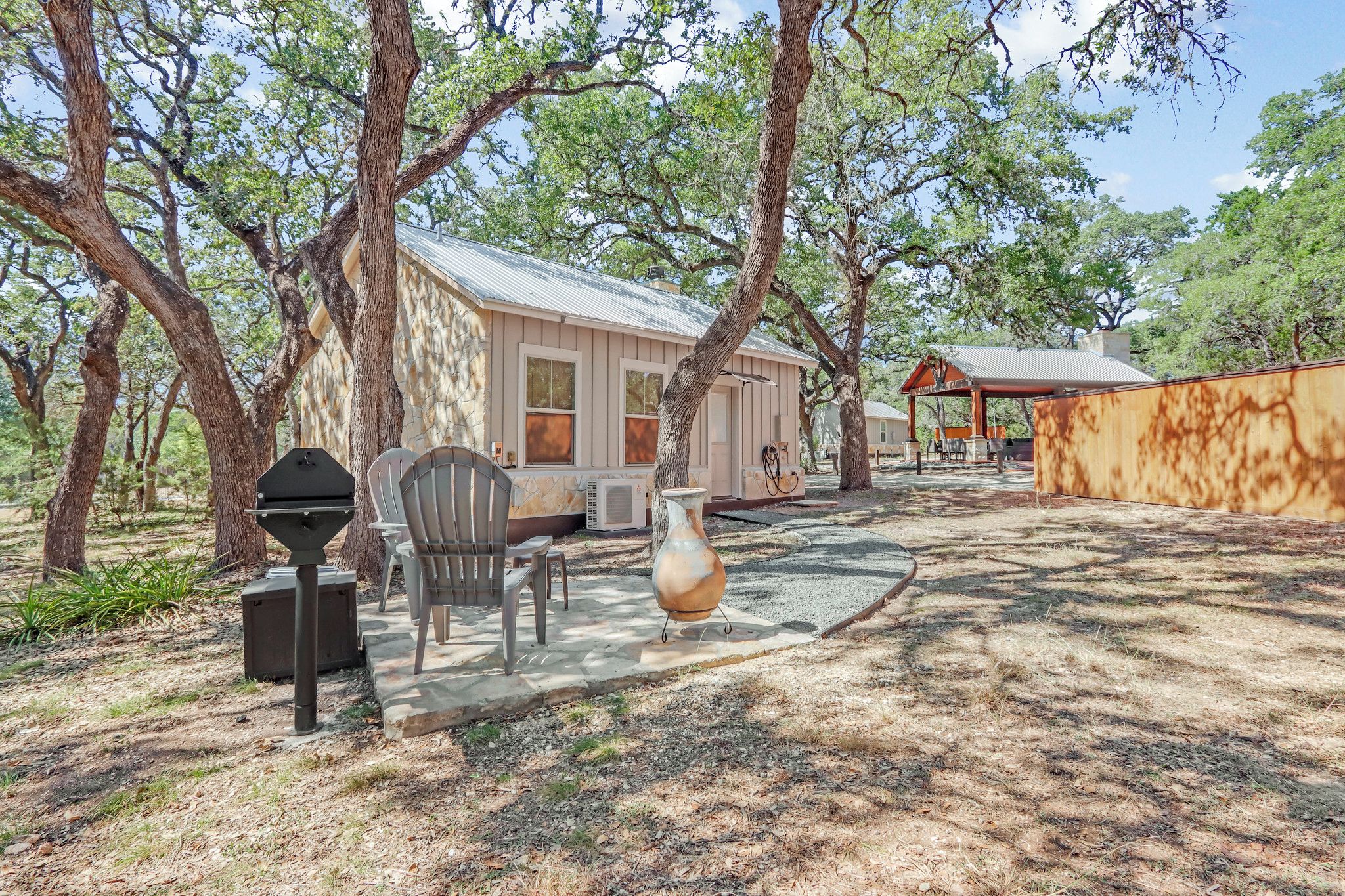 A rustic cabin with a metal roof, surrounded by trees. There is a covered outdoor seating area with a table and chairs.