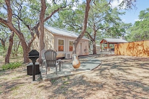A rustic cabin with a metal roof, surrounded by trees. There is a covered outdoor seating area with a table and chairs. - Texas Sage I Cabin close to the Square! (Wimberley)
