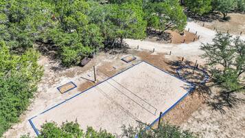 An aerial view of a sandy sports court with blue lines, surrounded by dense green trees.
