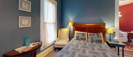 A hotel room with a bed, bedside lamps, a chair, a table with a vase and book, and a window with curtains.
