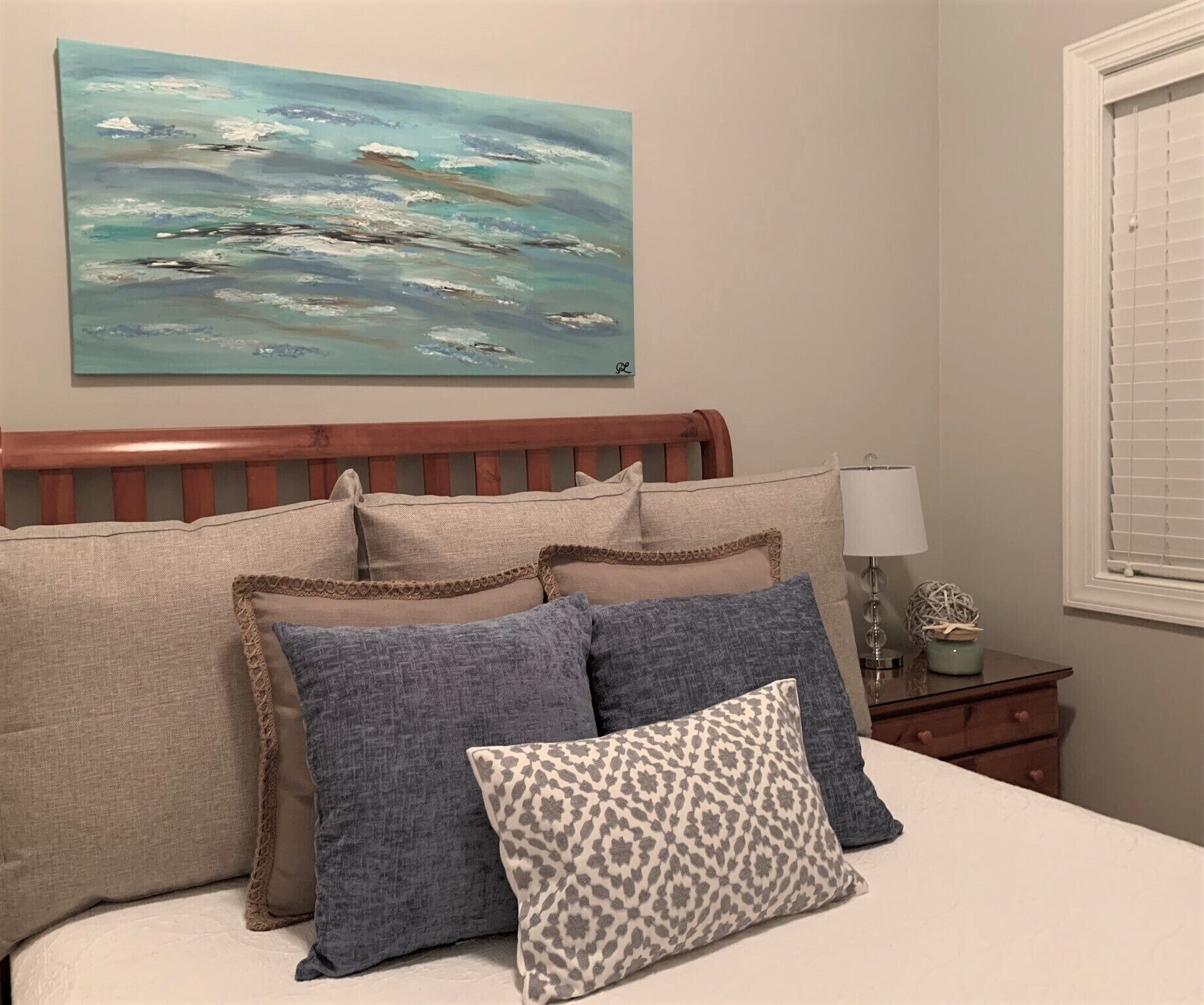A bedroom with a wooden headboard, a bed with multiple pillows, a painting of a seascape, a bedside lamp, and a window with blinds.