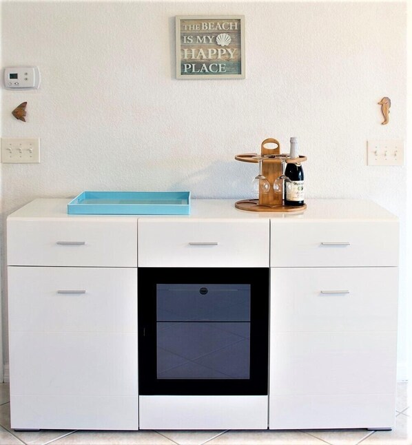Un escritorio blanco con cajones, un pequeño electrodoméstico negro, una botellera de madera con una botella y copas, y un cartel en la pared.