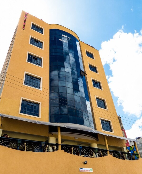 A multi-story building with a yellow facade and a glass elevator shaft. - The Fontage Hotel (Mlolongo)