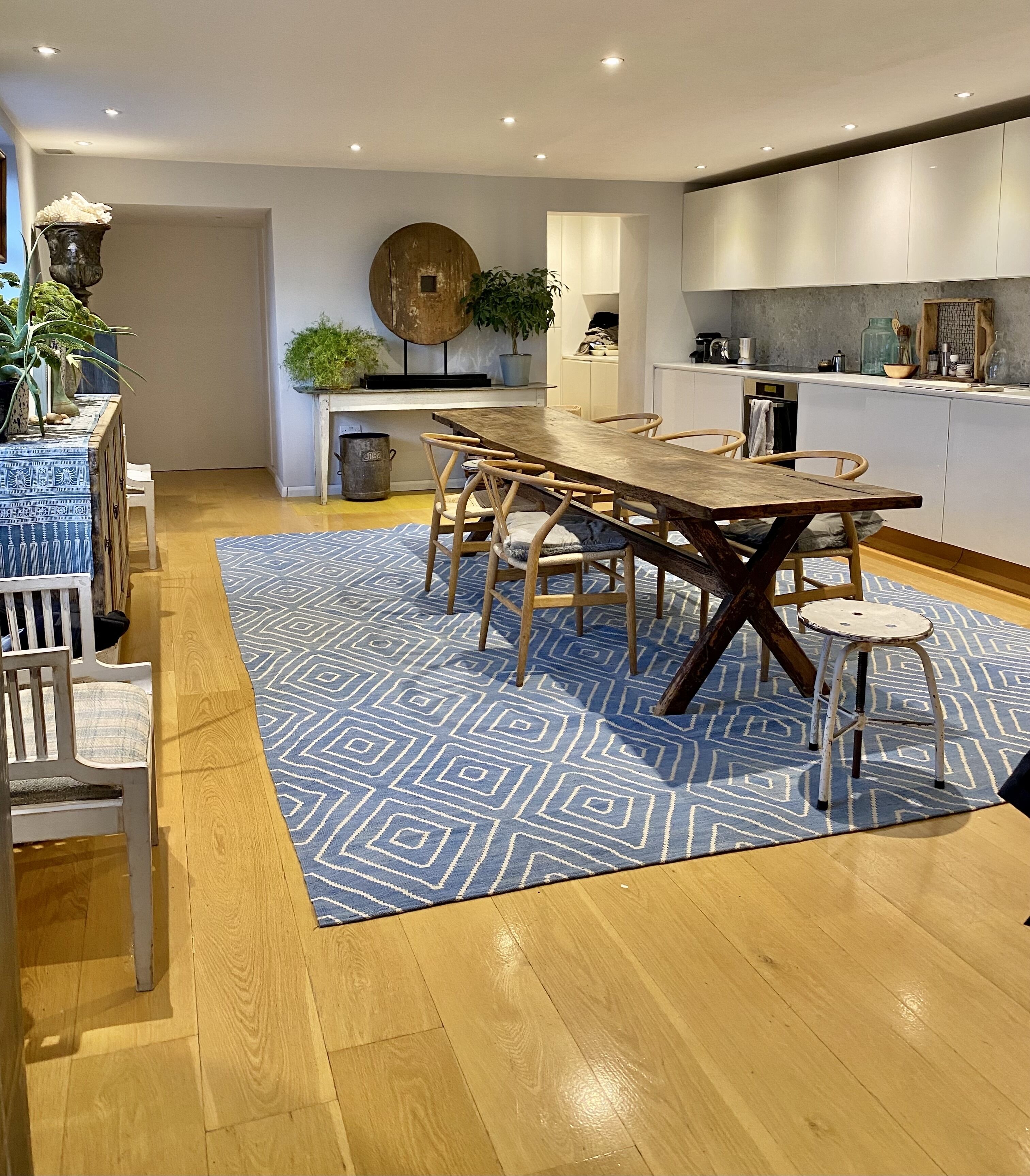 A modern kitchen with a wooden dining table, white cabinets, and a blue patterned rug.