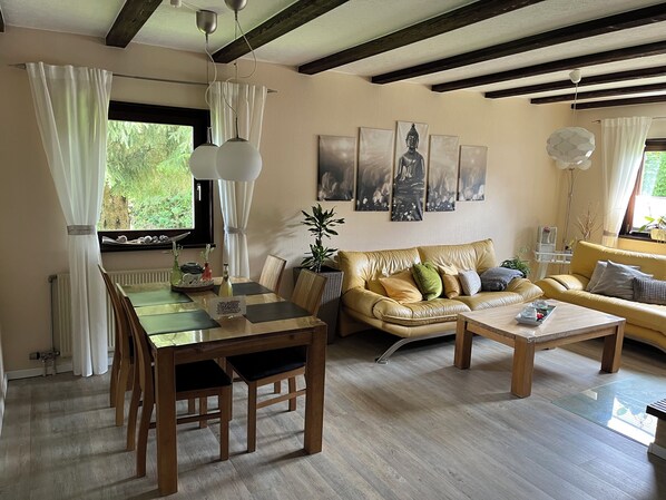 A living room with a wooden dining table, a sofa, and a coffee table. There are curtains, a plant, and artwork on the walls.