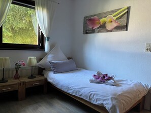A single bed with white bedding, a bedside table with lamps, a vase with flowers, and a window with curtains.
