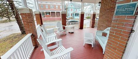 A covered porch with white Adirondack chairs, wicker furniture, and a brick wall with a historic register plaque.