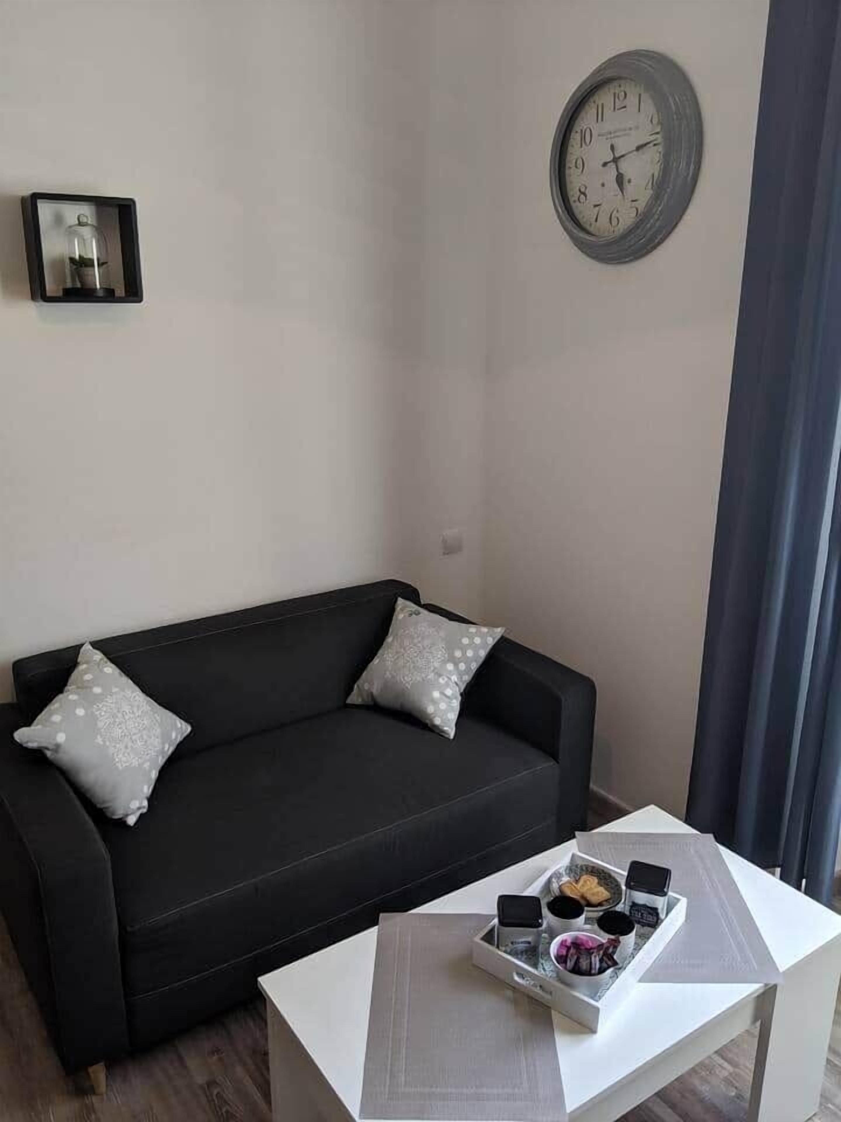 Un salón moderno con un sofá negro, dos almohadas grises, una mesa de centro blanca con una bandeja de aperitivos y un reloj de pared.
