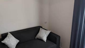 Un salón moderno con un sofá negro, dos almohadas grises, una mesa de centro blanca con una bandeja de aperitivos y un reloj de pared.