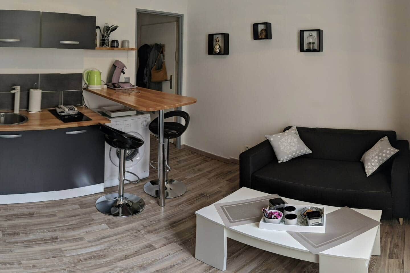 Una cocina moderna con encimera de madera, dos taburetes negros, una mesa de centro blanca con tazas y una caja, un sofá negro con cojines estampados y una pared con tres cuadros enmarcados.